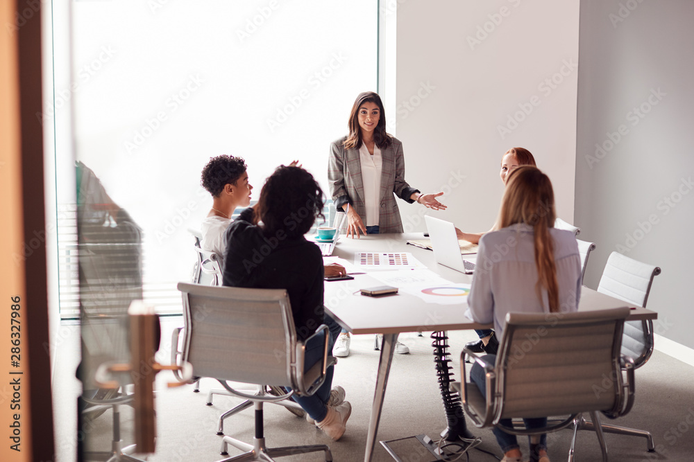 Female Boss Gives Presentation To Team Of Young Businesswomen Meeting Around Table In Modern Office